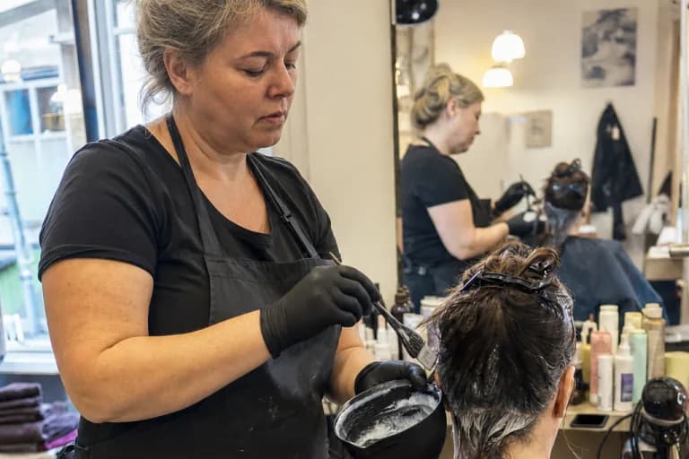 Dieselbe Friseurin im selben Salon arbeitet ruhig mit beiden Händen an der Kundin, das Telefon liegt dunkel auf der Ablage.